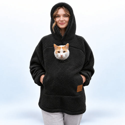 A woman wearing a black sherpa hoodie with a realistic cat face printed on the front pocket, paired with light gray sweatpants, against a light blue studio background.