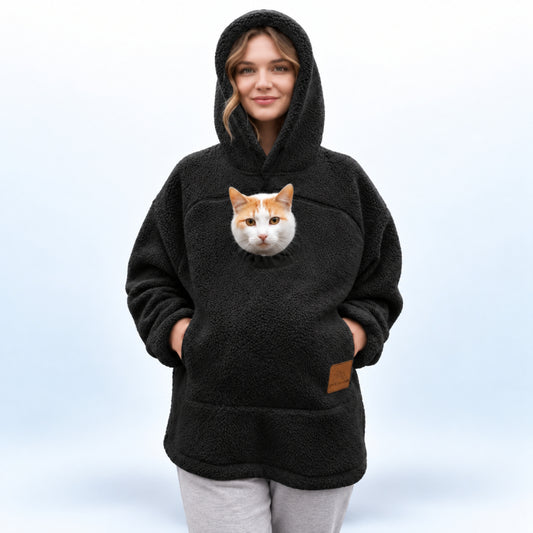 A woman wearing a black sherpa hoodie with a realistic cat face printed on the front pocket, paired with light gray sweatpants, against a light blue studio background.